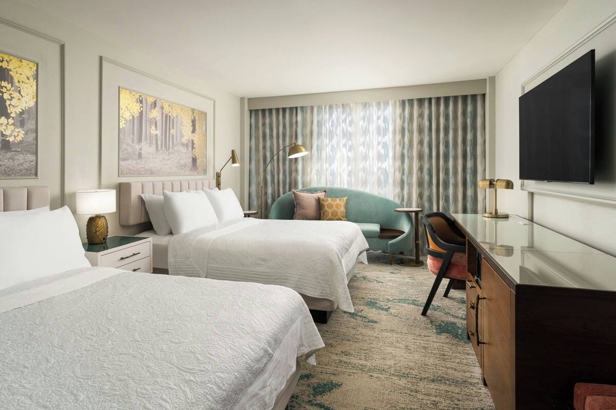 A hotel room with two beds, a desk, a chair, a television, and a large window with curtains.