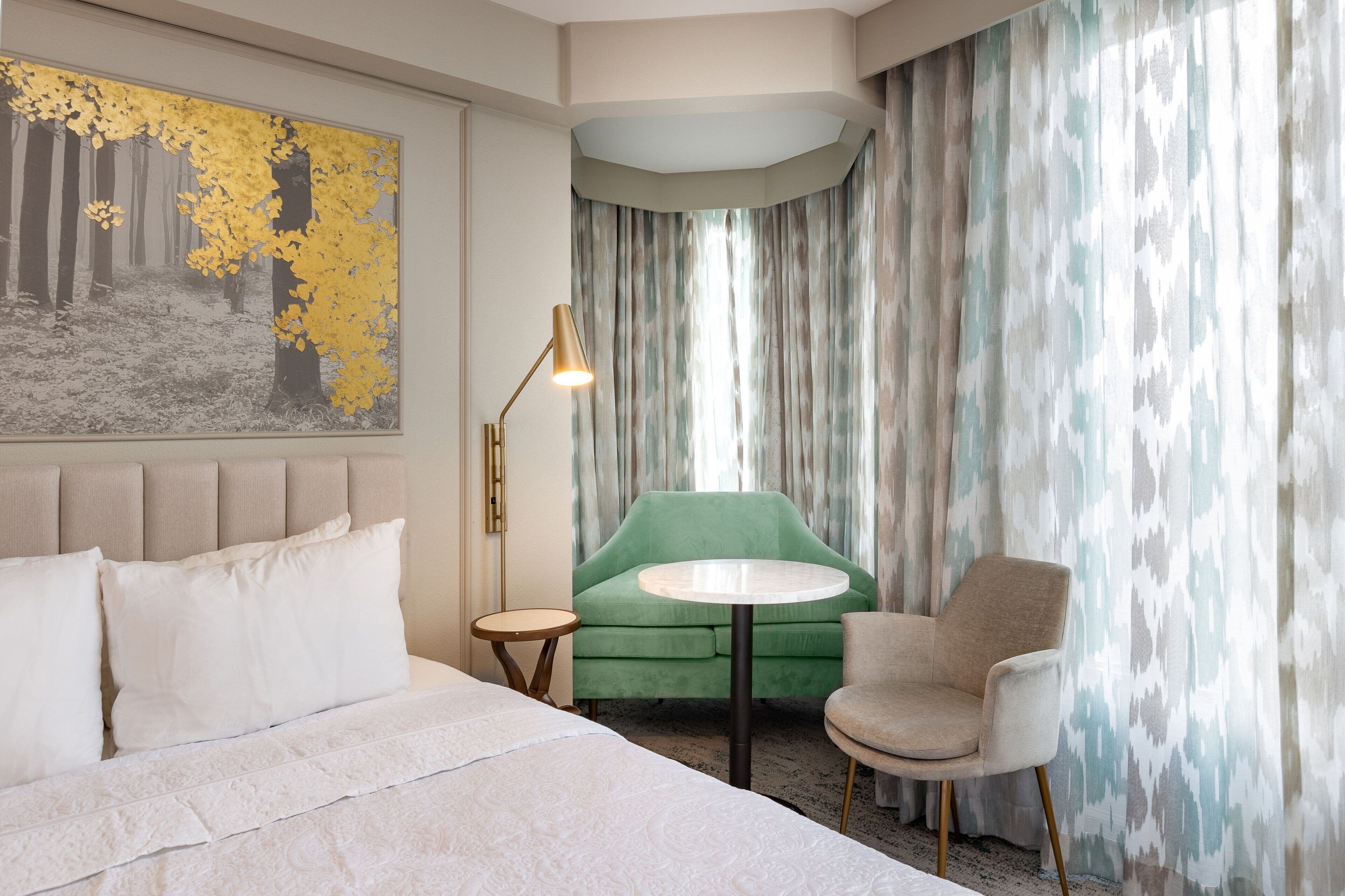 Une chambre d’hôtel avec un grand lit, un fauteuil vert, une table ronde et un tableau représentant une forêt.