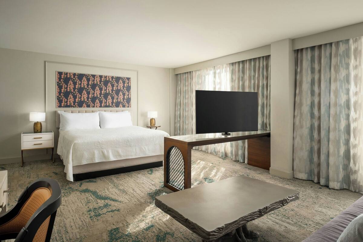 A hotel room with a bed, a TV, a desk, a chair, and a large window with curtains.