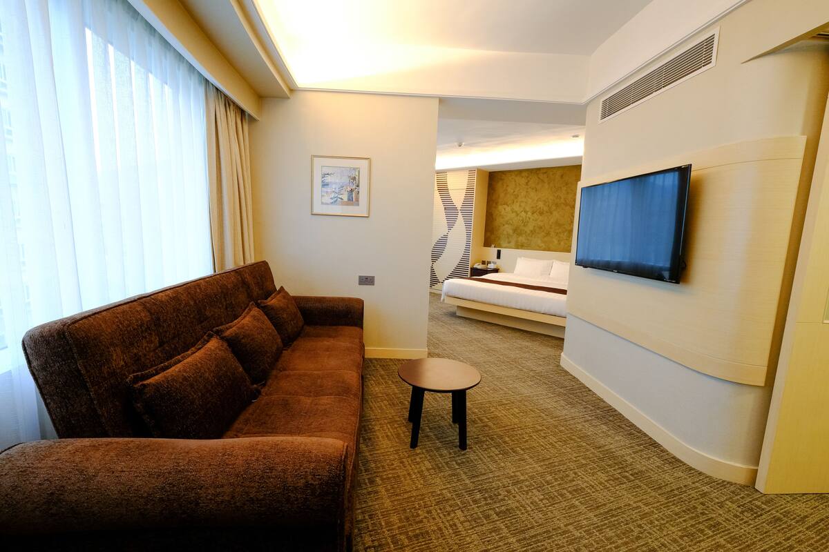 Een hotelkamer met een bruine bank, een klein rond tafeltje, een televisie aan de muur en een bed op de achtergrond.