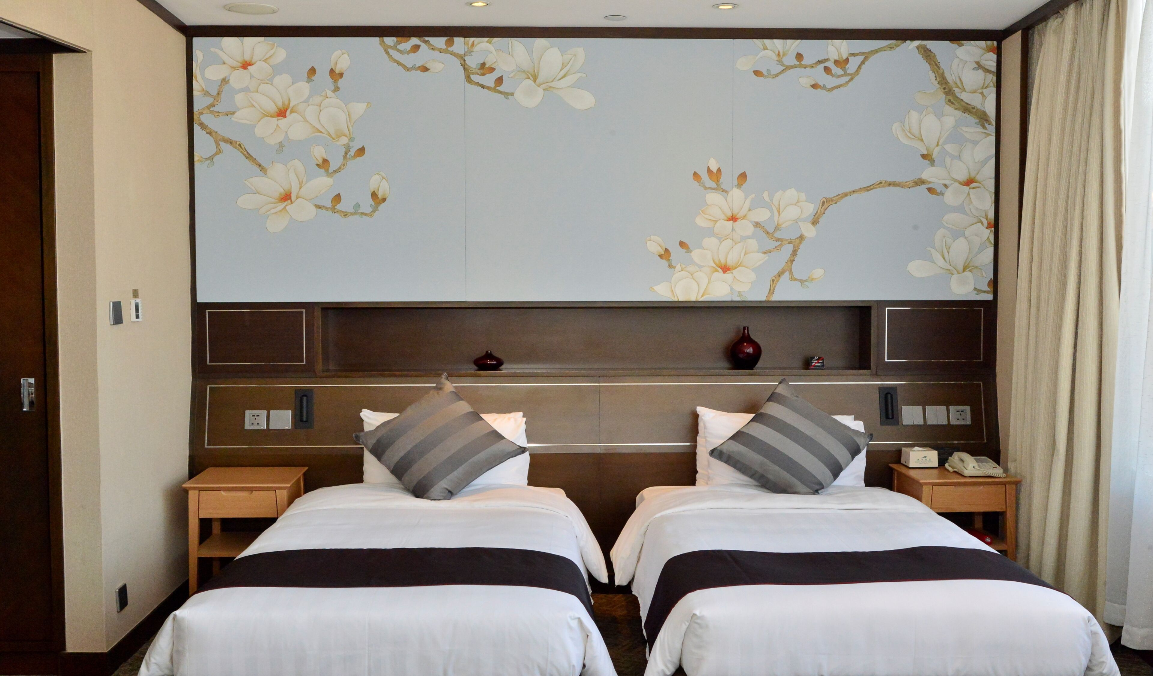 Une chambre d’hôtel avec deux lits, une grande fresque murale à motifs floraux, une tête de lit en bois et une table de chevet avec un téléphone.