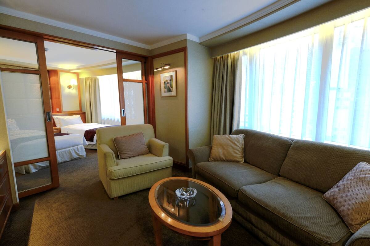 Een hotelkamer met een bed, een bank, een stoel en een kleine tafel. Er is een raam met gordijnen en uitzicht op gebouwen.