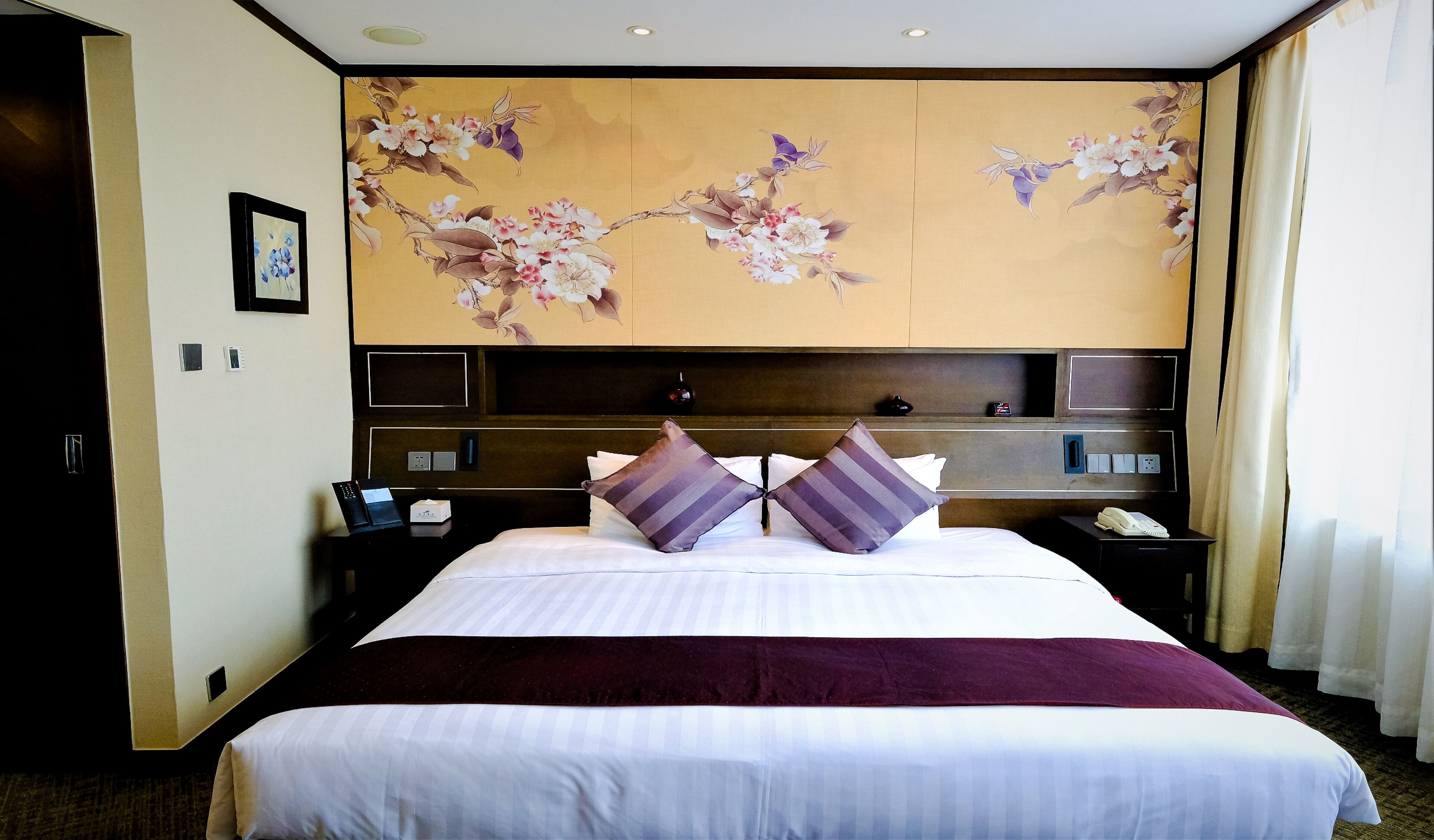 A hotel room with a large bed, a headboard with a floral mural, a bedside table with a phone, and a nightstand with a lamp.