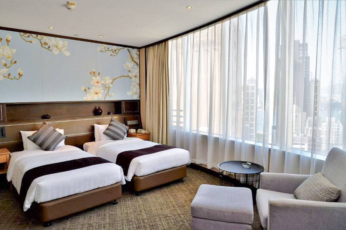 Een hotelkamer met twee bedden, een bank, een klein tafeltje en een groot raam met gordijnen.