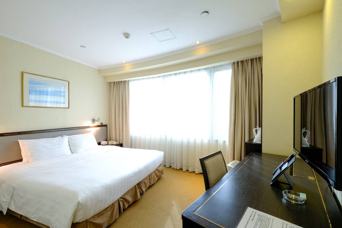 Een hotelkamer met een groot bed, een bureau met een flatscreen televisie, een raam met gordijnen en een ingelijst kunstwerk aan de muur.