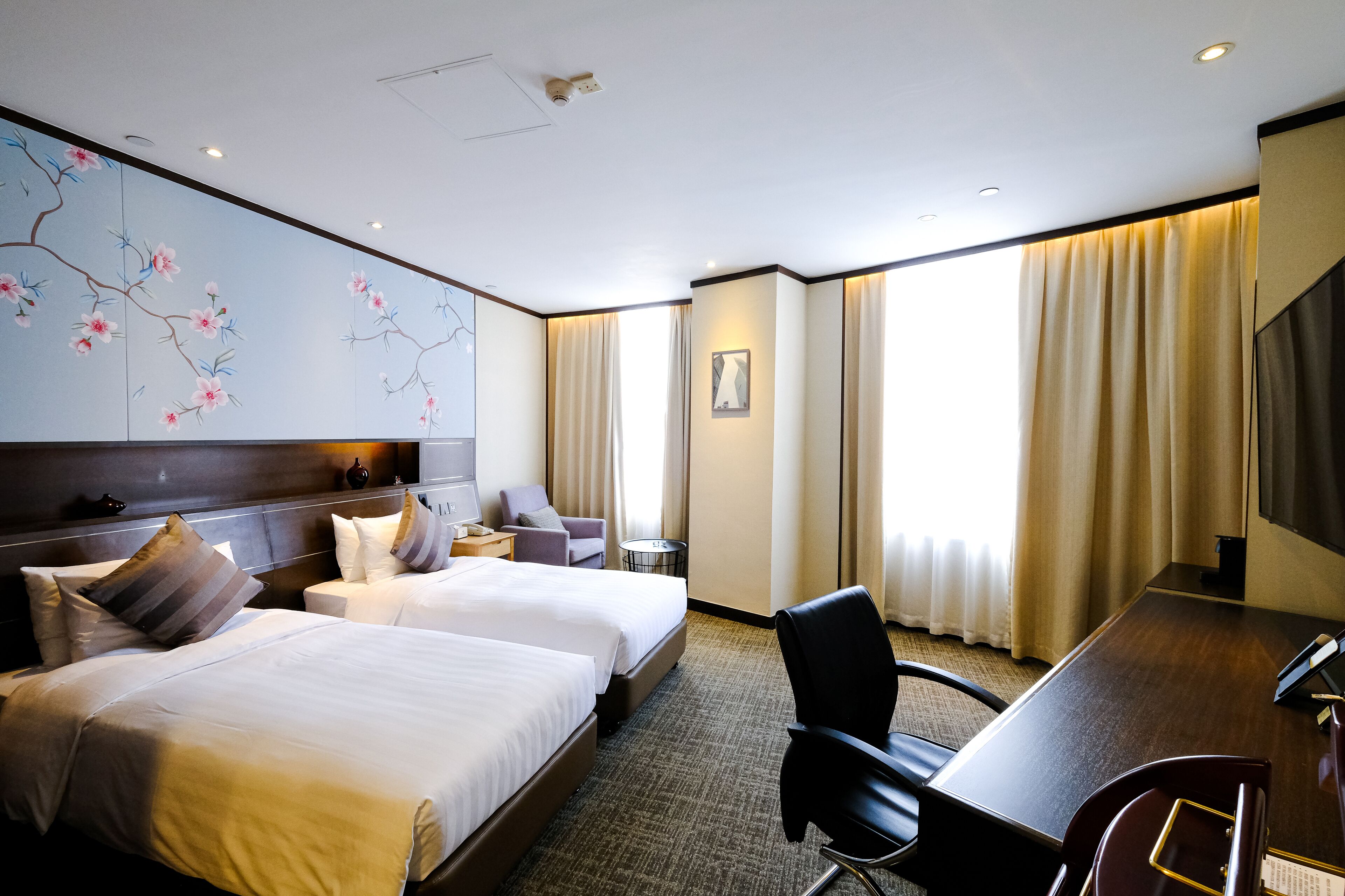 Ein Hotelzimmer mit zwei Betten, einem Schreibtisch, einem Stuhl, einem kleinen Tisch und einem großen Fenster mit Vorhängen.