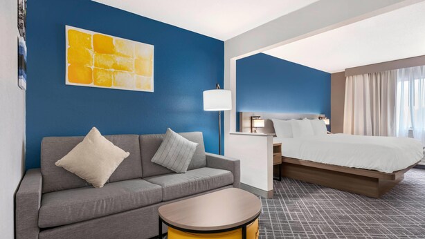 A hotel room with a grey sofa, a yellow side table, a bed with white bedding, a nightstand, a lamp, and a blue accent wall with a yellow artwork. - Comfort Inn & Suites Wisconsin Dells (Wisconsin Dells)