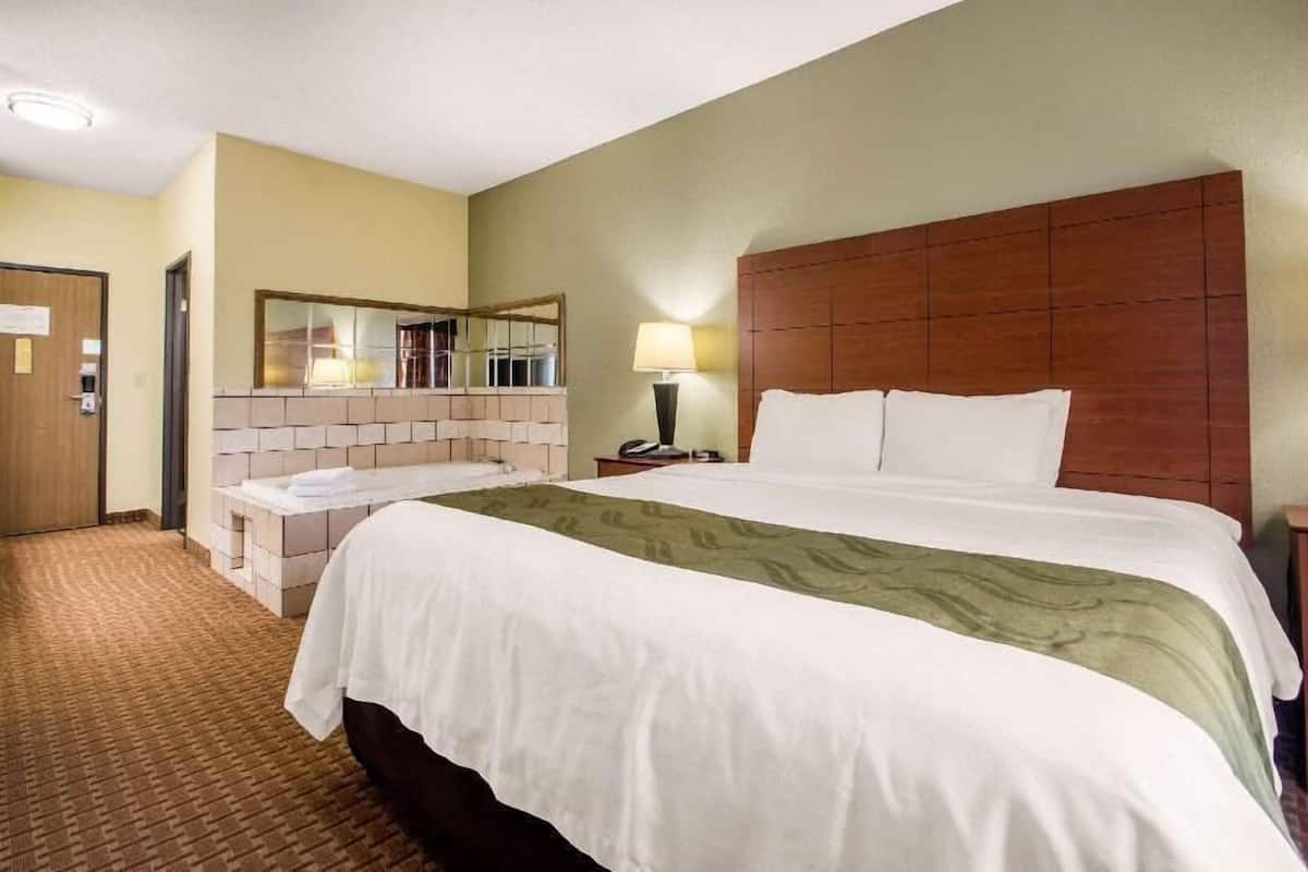A hotel room with a large bed, a small bedside table, a nightstand, a mirror, and a bathroom with a sink and a towel rack.