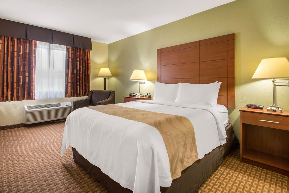A hotel room with a bed, two bedside tables with lamps, a chair, an air conditioning unit, and a window with curtains.
