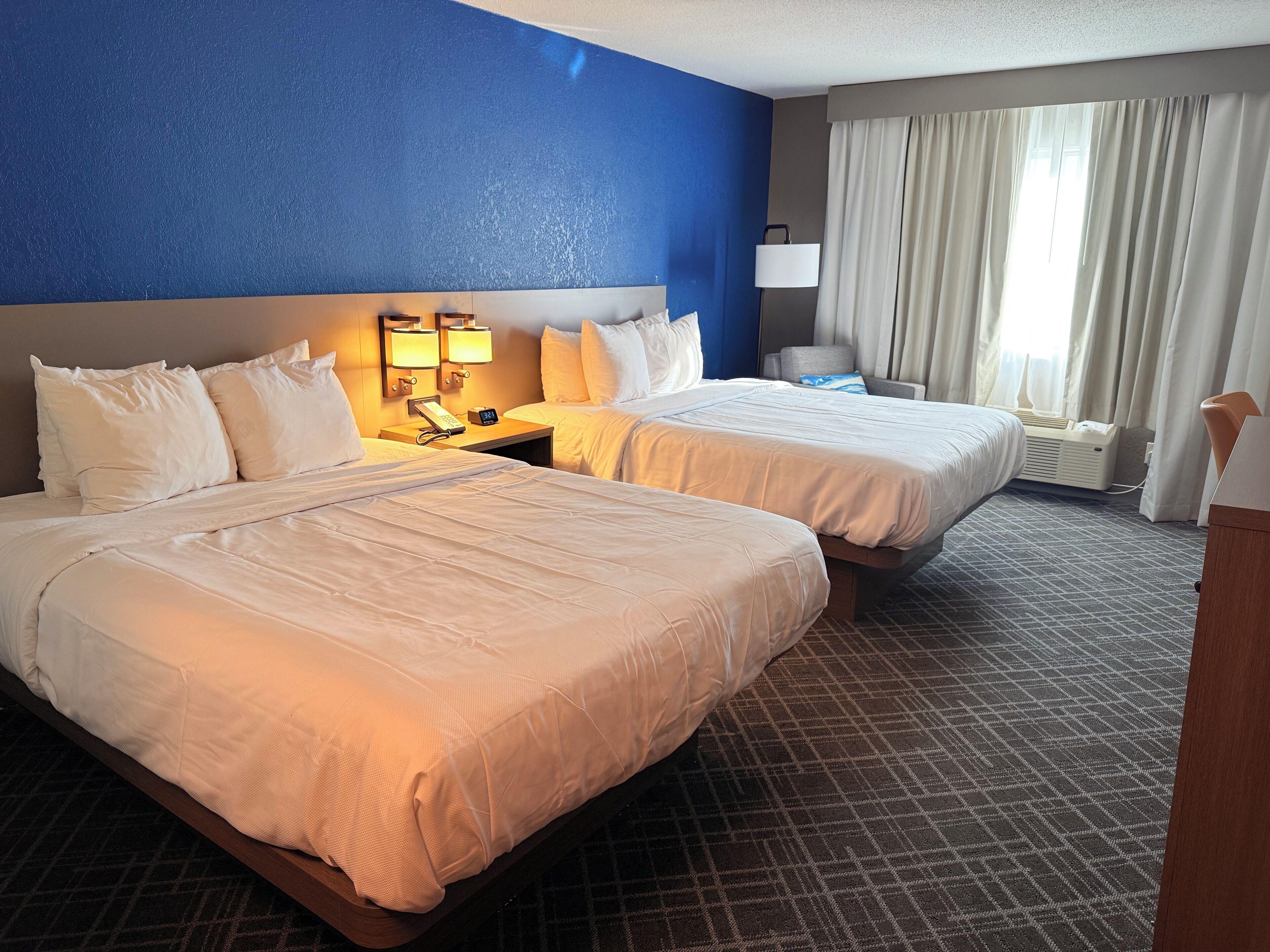 A hotel room with two beds, a nightstand with a phone and lamp, a window with curtains, and a carpeted floor.