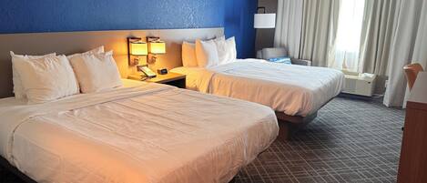 A hotel room with two beds, a nightstand with a phone and lamp, a window with curtains, and a carpeted floor.