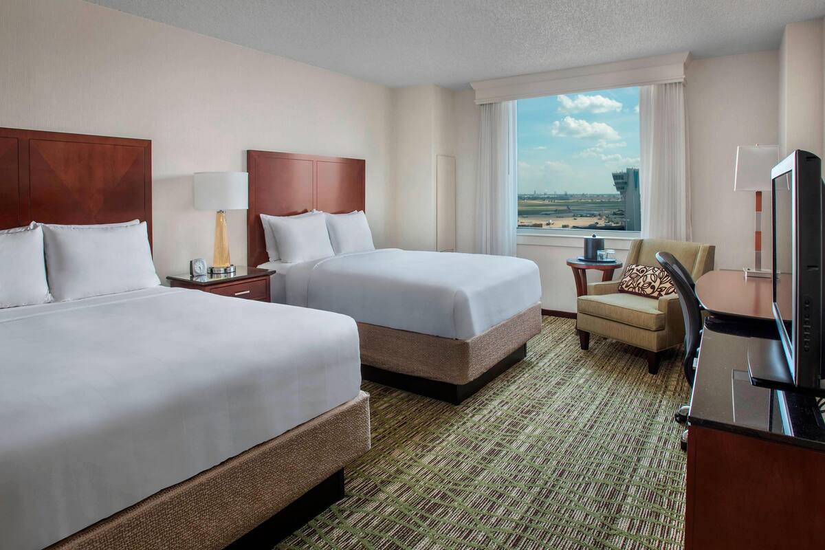 Habitación de hotel con dos camas, un escritorio con televisor, una silla y ventana con vista a la ciudad.