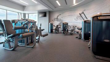 A modern gym with various exercise machines, large windows, and a wall-mounted TV.