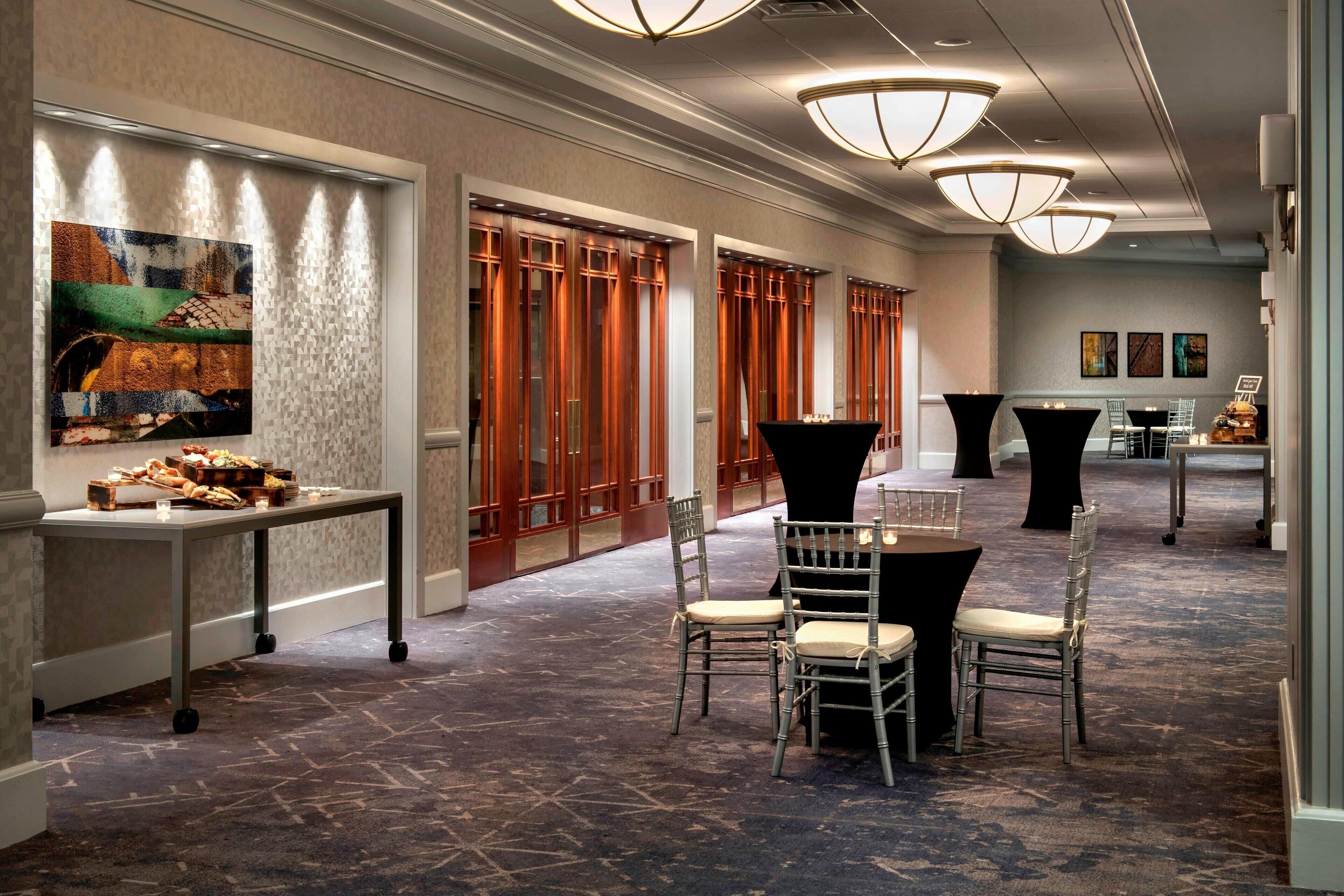 A hotel corridor with a carpeted floor, a table with food, chairs, and a painting on the wall.