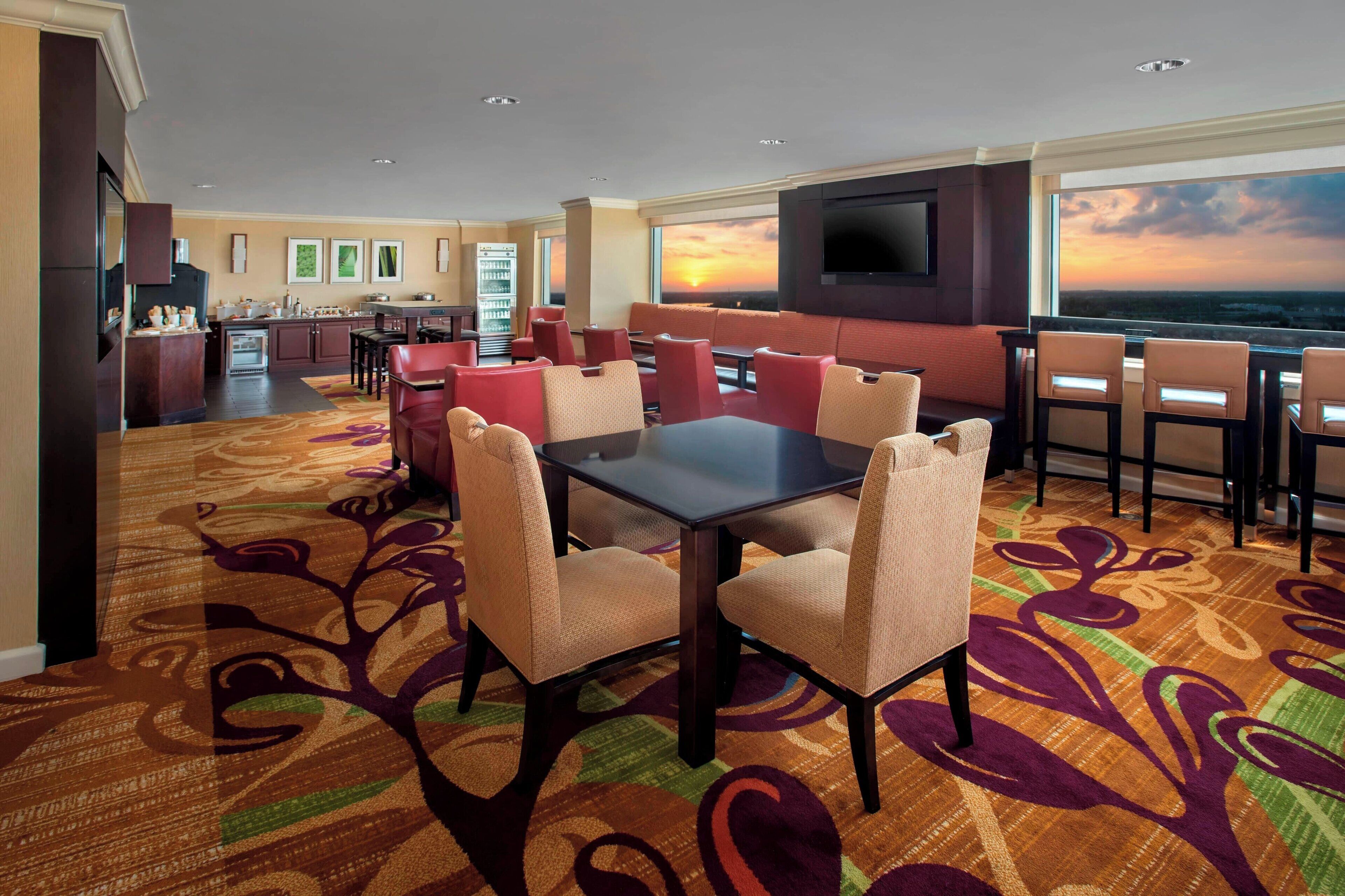 A hotel room with a dining area, a kitchen, and a large window with a sunset view.