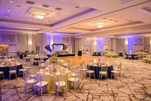 Un gran salón de baile con mesas redondas y sillas, un escenario con un arreglo floral y una pista de baile.