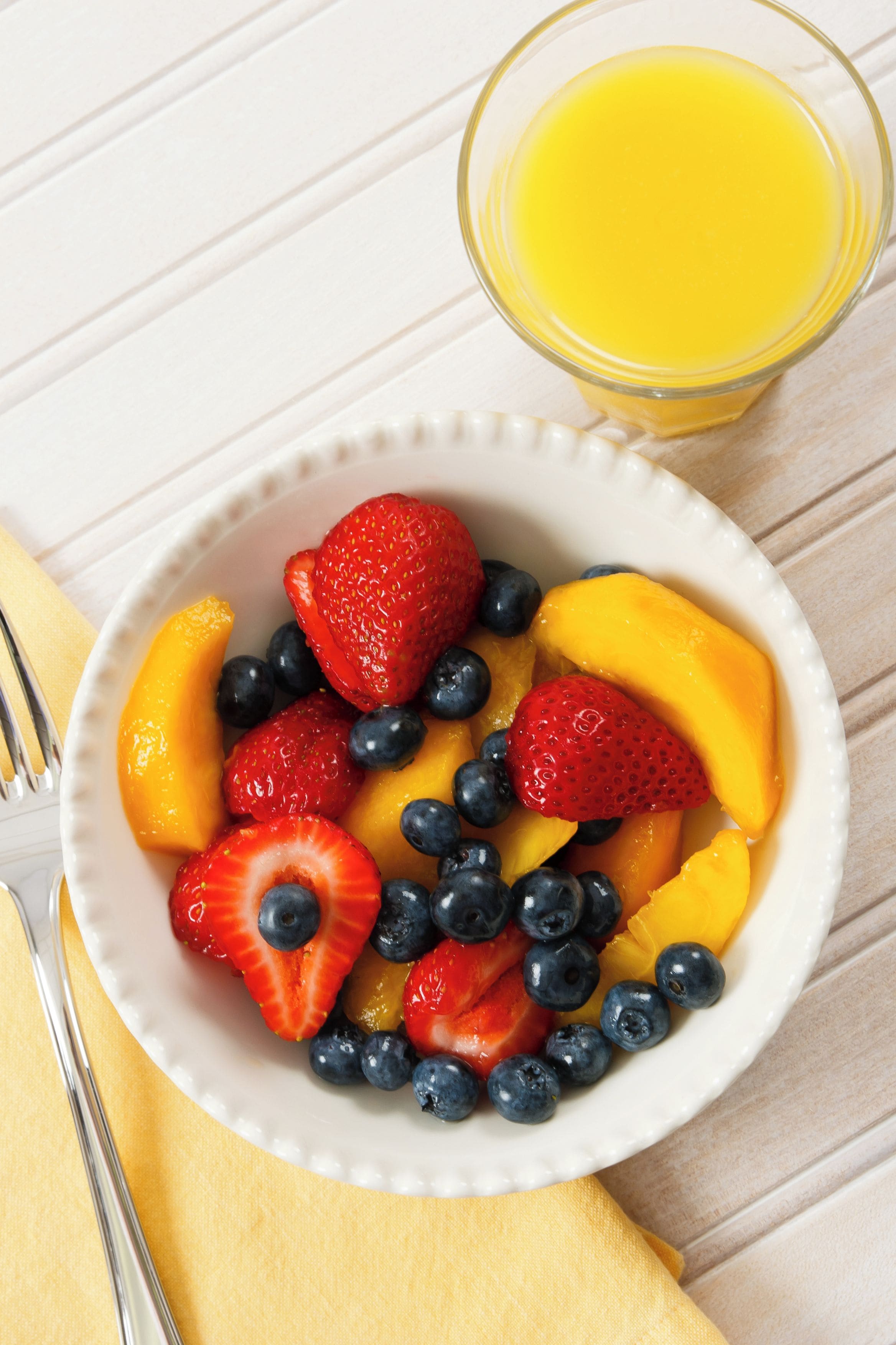 Un tazón de fruta con fresas, arándanos y melocotones, junto a un vaso de jugo de naranja sobre una superficie blanca.