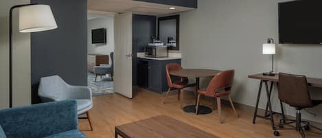 A modern hotel room with a living area, a dining table, a TV mounted on the wall, and a kitchenette.