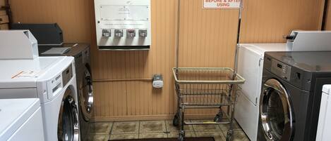 A laundry facility with washers and dryers, a soap and bleach softener dispenser, and a sign instructing to check and clean the dryer vent.