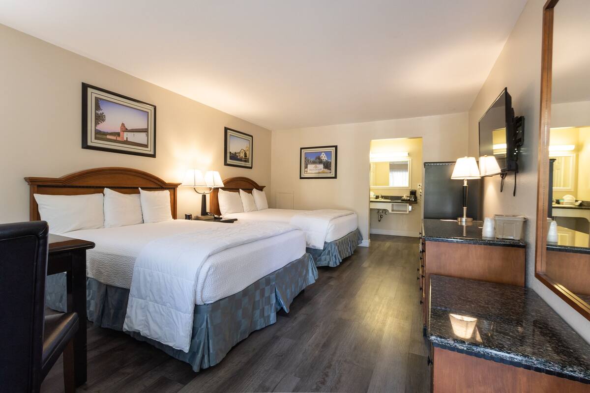 A hotel room with two beds, a desk, a chair, a vanity with a mirror, and a TV.