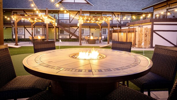 Un área de estar al aire libre circular con una chimenea, rodeada de sillones de mimbre, bajo un porche cubierto con luces colgantes.