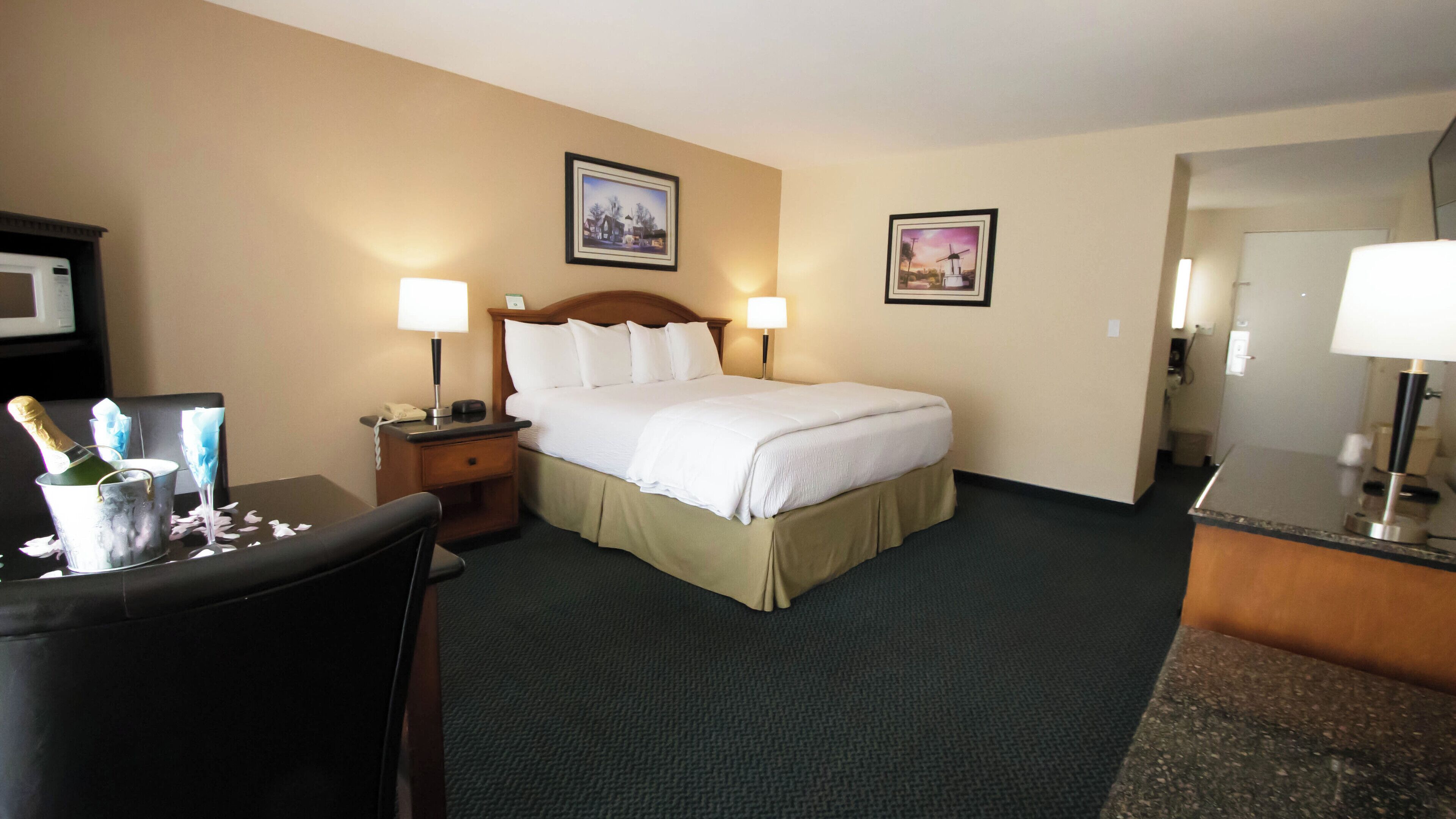 A hotel room with a bed, two bedside lamps, a nightstand with a phone, a chair, a desk with a lamp, and a minibar.