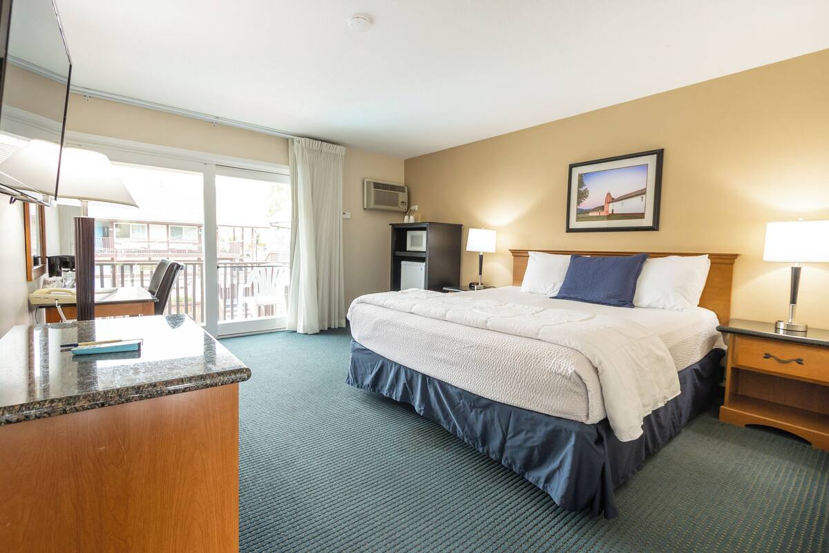 A hotel room with a bed, a desk, a chair, a lamp, a picture on the wall, and a view of the outside.