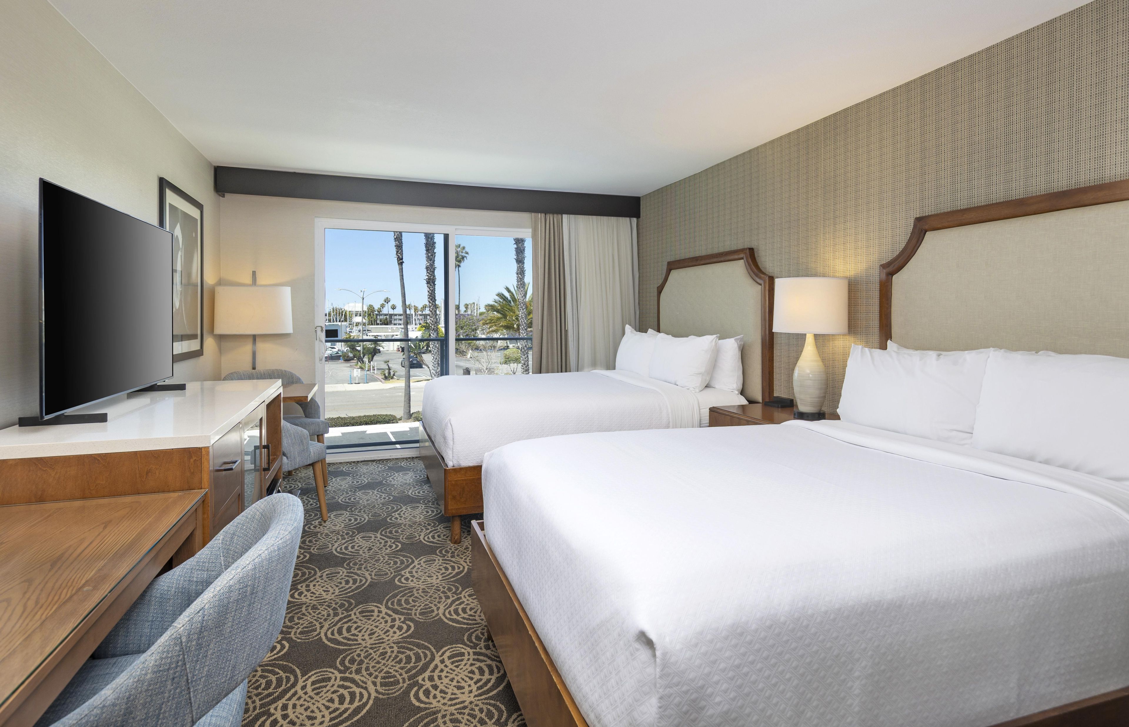 Habitación de hotel con dos camas, un escritorio, televisión y vistas a palmeras y edificios.