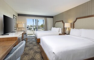 Habitación de hotel con dos camas, un escritorio, televisión y vistas a palmeras y edificios.