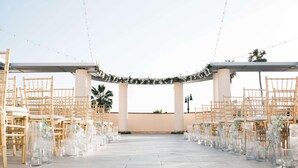 An outdoor wedding venue with rows of bamboo chairs and glass tables, decorated with greenery and string lights. - Redondo Beach Hotel, Tapestry Collection by Hilton (Redondo Beach)