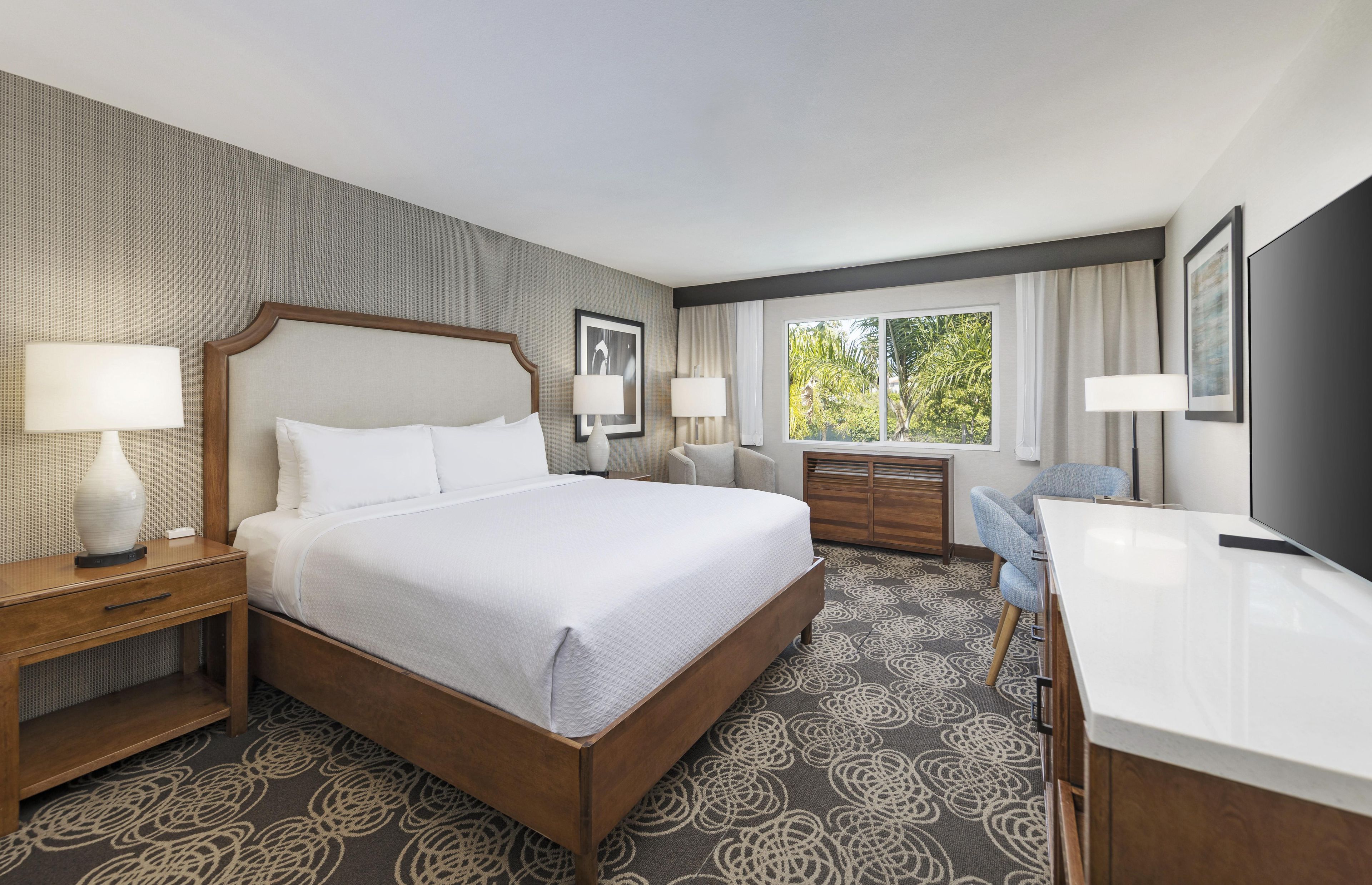 A hotel room with a large bed, bedside tables, a desk, and a window with a view of trees.