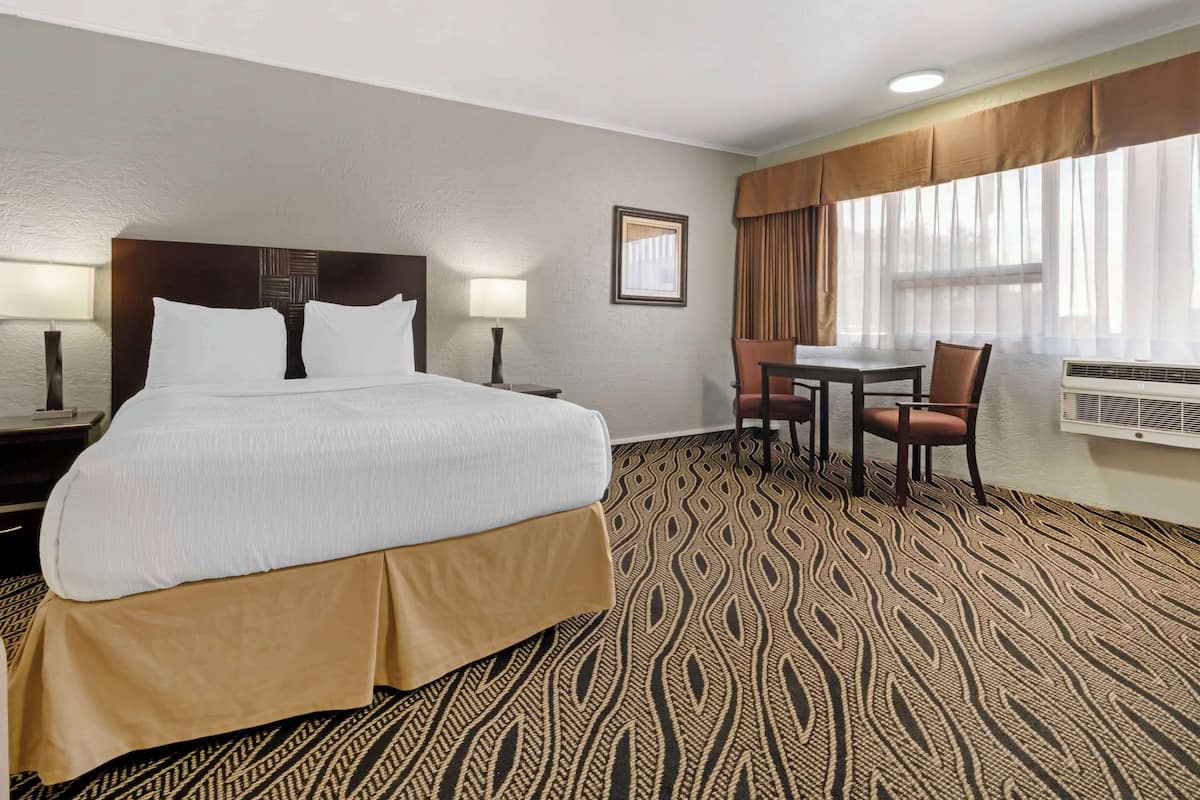 A hotel room with a large bed, a desk, two chairs, a window with curtains, and an air conditioning unit.
