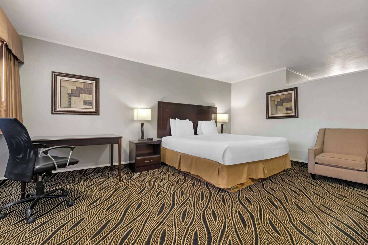 A hotel room with a large bed, a desk with a chair, a lamp, and two framed pictures on the walls.