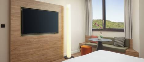 Habitación de hotel con una cama grande, un televisor de pantalla plana montado en un panel de madera, una mesita redonda con un florero, un sofá verde y una ventana con vista a follaje.