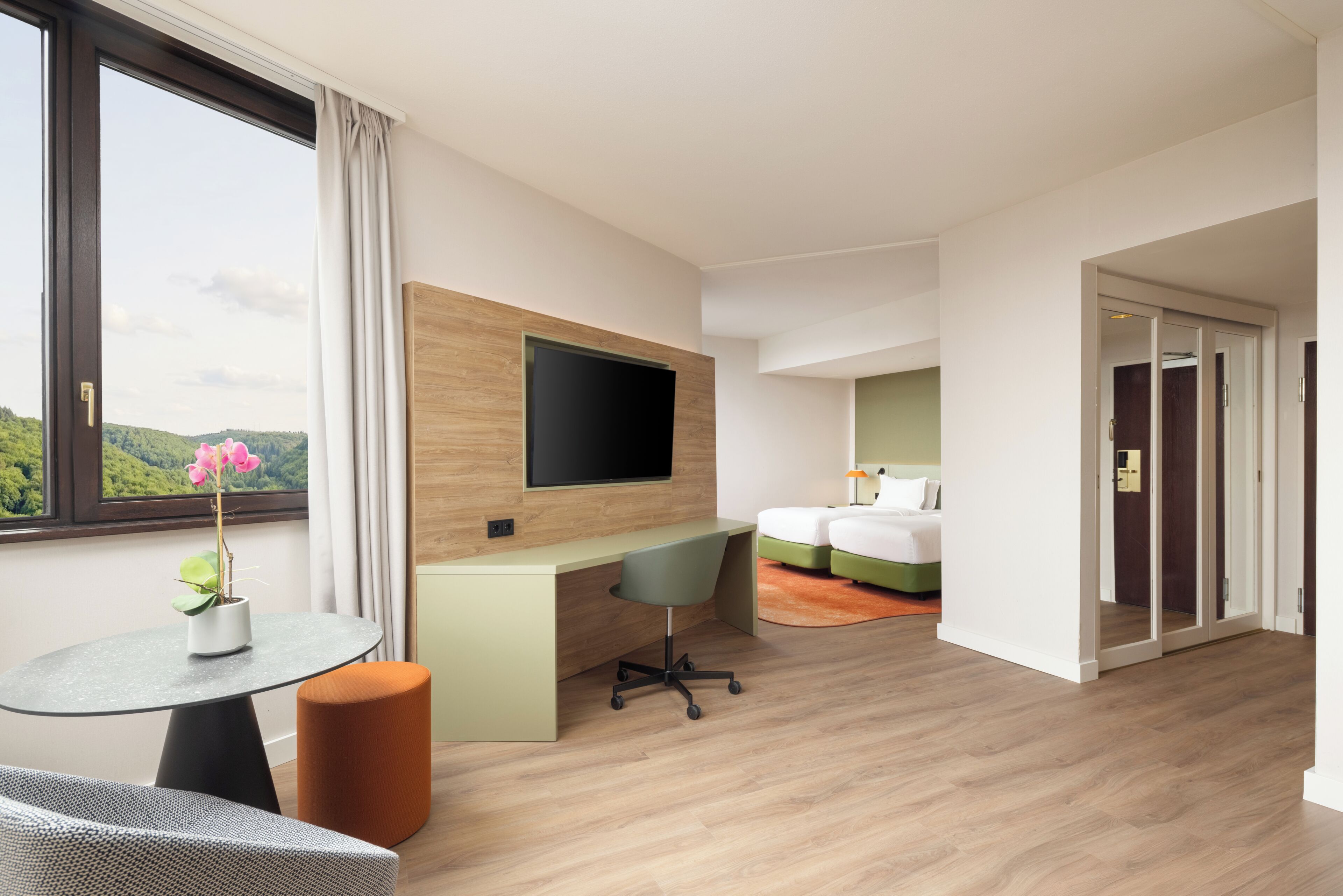 Hotellihuone, jossa on sänky, työpöytä, televisio, ikkuna verhoilla ja näkymä vehreään maisemaan.