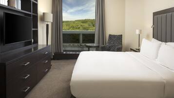 A hotel room with a bed, a TV, a desk, and a window with a view of greenery.