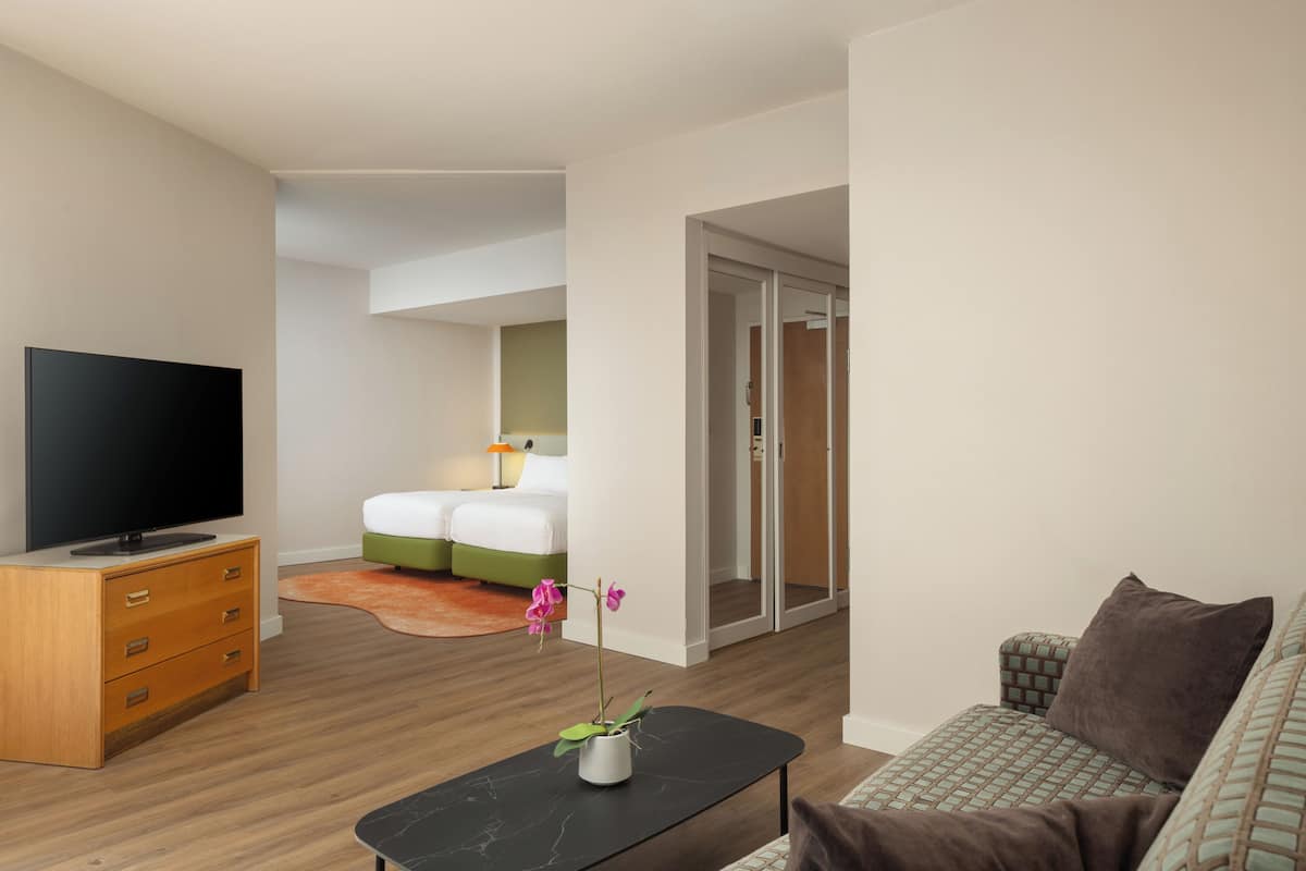 Moderni hotellihuone, jossa on taulutelevisio, sohva, vihreillä tekstiileillä varustettu sänky, pieni pöytä maljakkoineen ja vaatehuone.