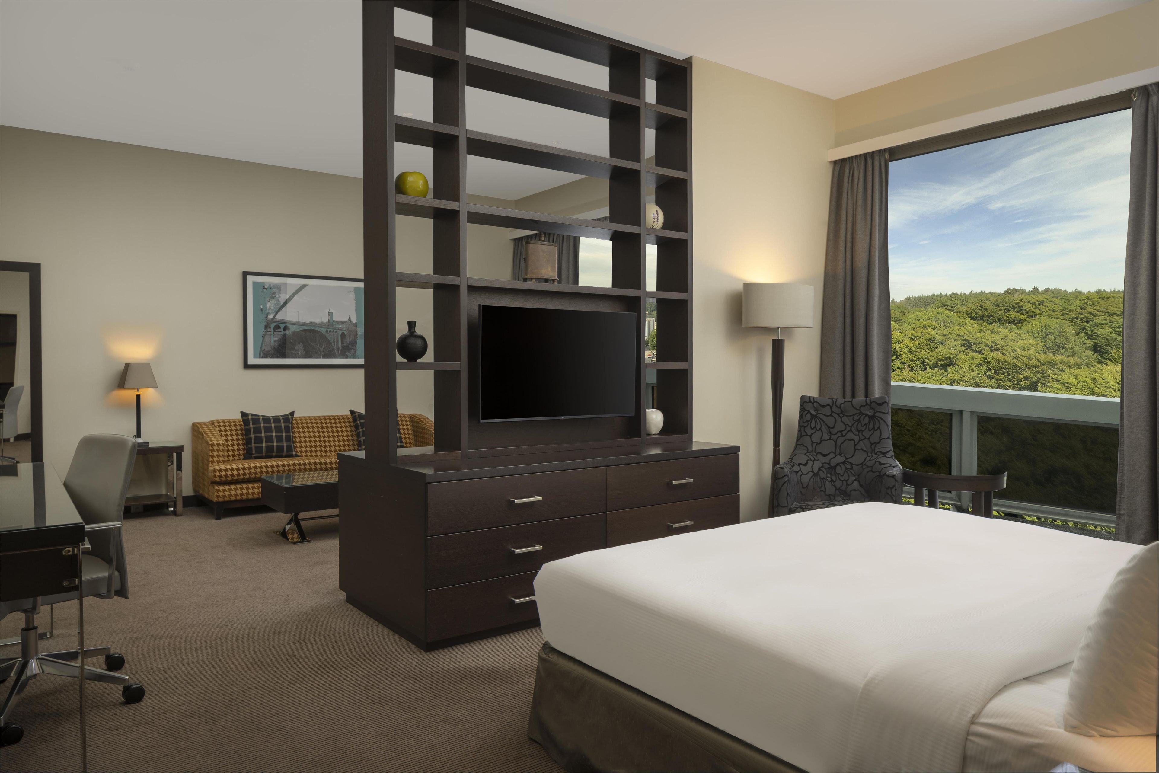 A hotel room with a large bed, a TV on a dark wooden stand, a desk with a chair, a sofa, and a large window offering a view of greenery.
