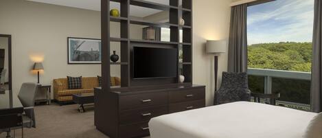 A hotel room with a large bed, a TV on a dark wooden stand, a desk with a chair, a sofa, and a large window offering a view of greenery.