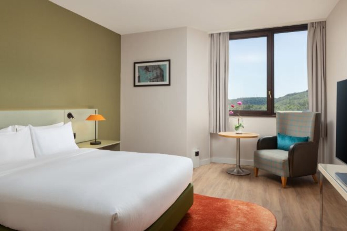 A hotel room with a bed, a chair, a small table, and a window with a view.