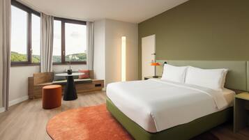 Een moderne hotelkamer met een groot bed, een zithoek met een bank en een poef, een nachtkastje met een lamp, en uitzicht op het omliggende landschap door grote ramen.