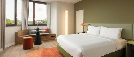 A modern hotel room with a large bed, a seating area with a sofa and ottoman, a nightstand with a lamp, and a view of the outdoors through large windows.