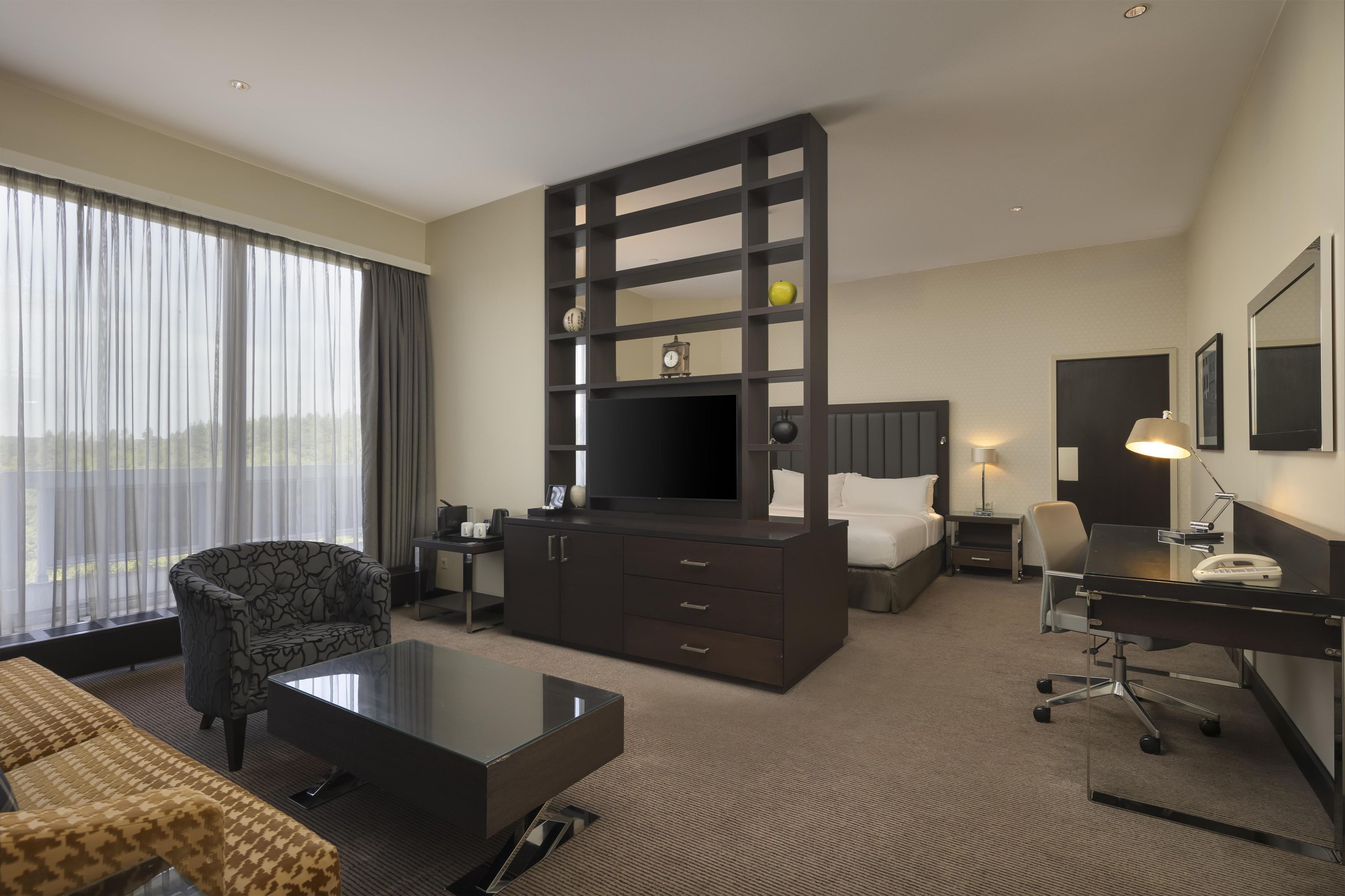 A hotel room with a large bed, a desk with a chair, a sofa, a television, and a window with curtains.