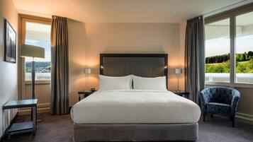 Habitación de hotel con una cama grande, dos mesitas de noche, una silla y vistas al exterior.