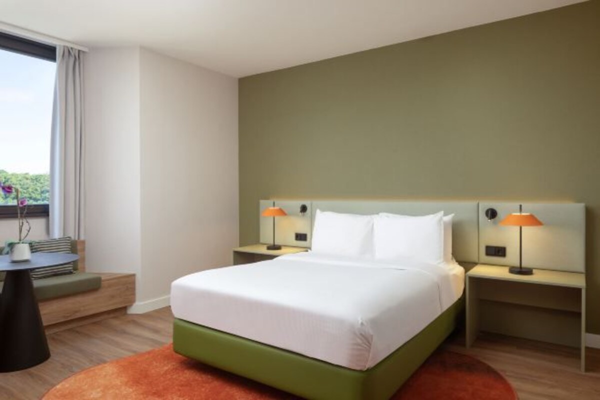 A hotel room with a bed, two bedside tables, a window with curtains, and a view of greenery outside.