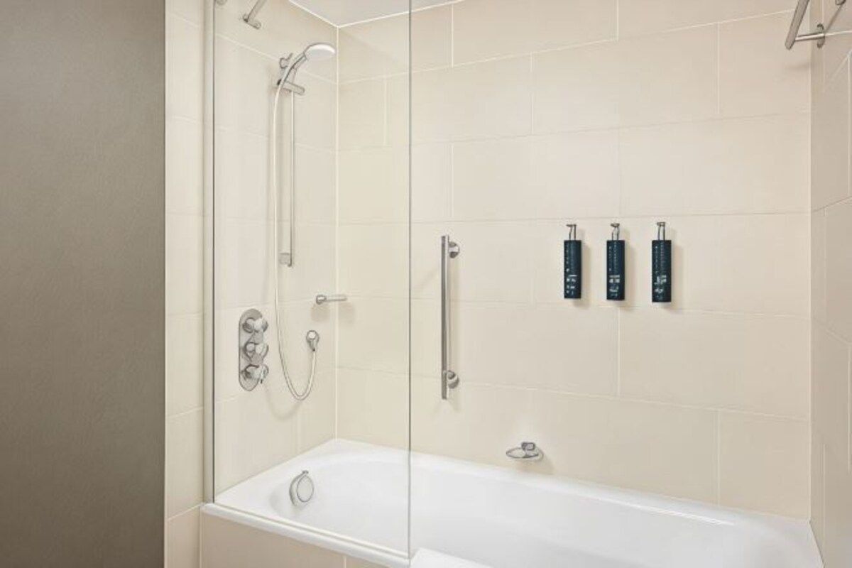 A bathroom with a glass-enclosed shower, a white bathtub, and three black toiletries on the wall.