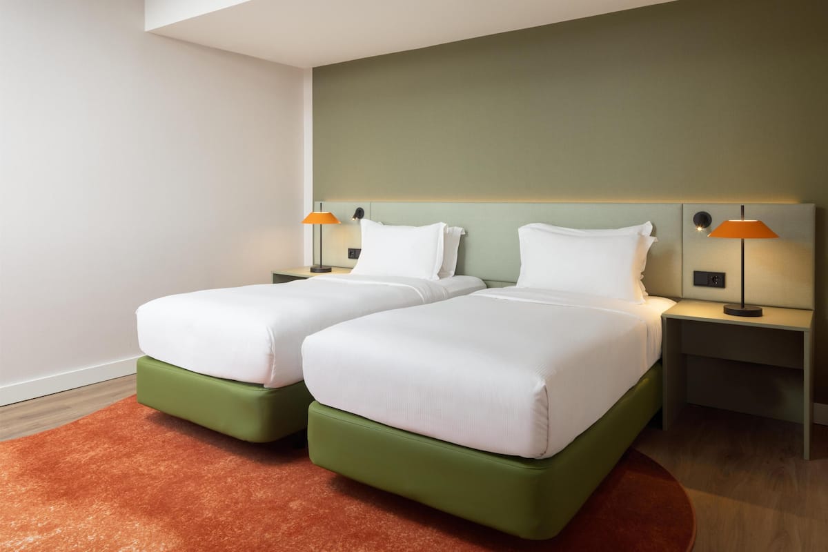 A hotel room with two beds, green headboards, white bedding, and two bedside lamps.