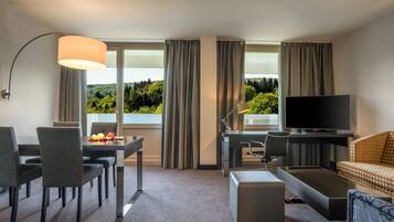 Habitación de hotel con mesa de comedor, sillas, sofá, televisor y una ventana grande con cortinas.