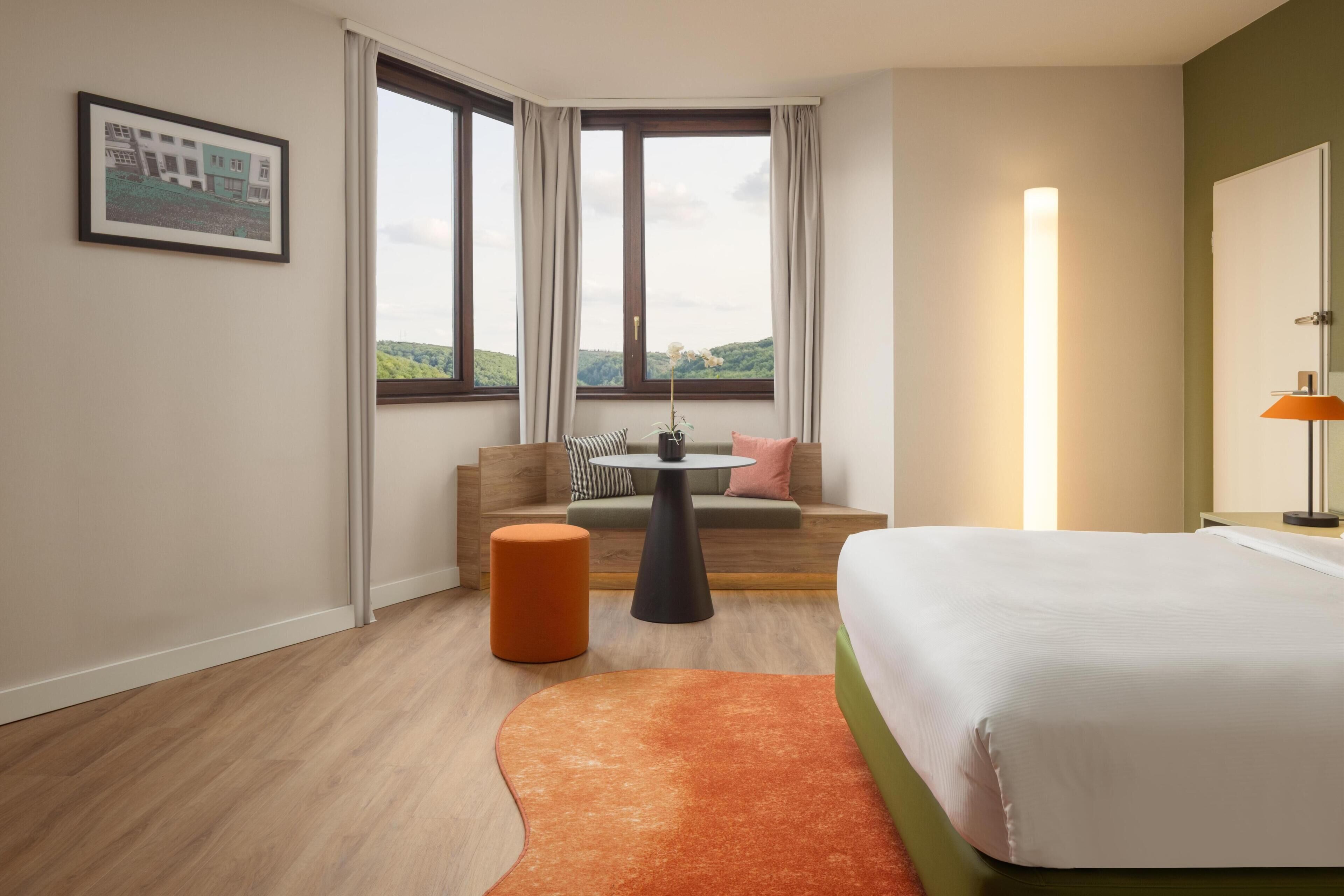 A hotel room with a bed, a sofa, a small table, a round orange ottoman, and a window with curtains.