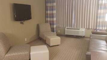 A hotel room with a flat-screen TV, patterned chairs, a sofa, a small ottoman, and a standing heater.